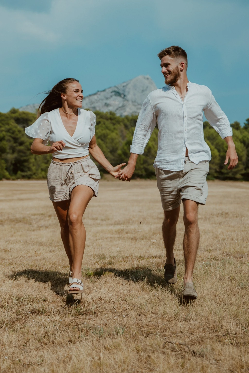 Virginie et Théo marchent main dans la main en souriant, avec la Sainte-Victoire en arrière-plan.